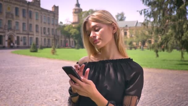 Pretty caucasian blonde female is swiping her phone and using it while standing on the street relaxed - Séquence, vidéo