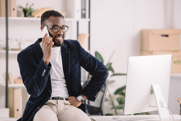bem sucedido empresário afro-americano falando smartphone enquanto sentado na mesa no escritório moderno com computador
 - Foto, Imagem
