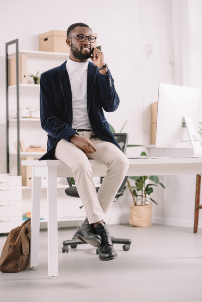 handsome successful african american businessman talking smartphone on while sitting on table in modern office   - Foto, afbeelding