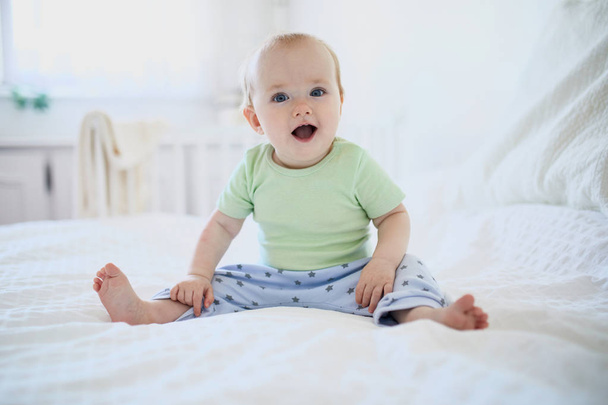 Baby girl sitting on bed and laughing. Happy healthy little child at home. Infant kid in sunny nursery - Photo, image