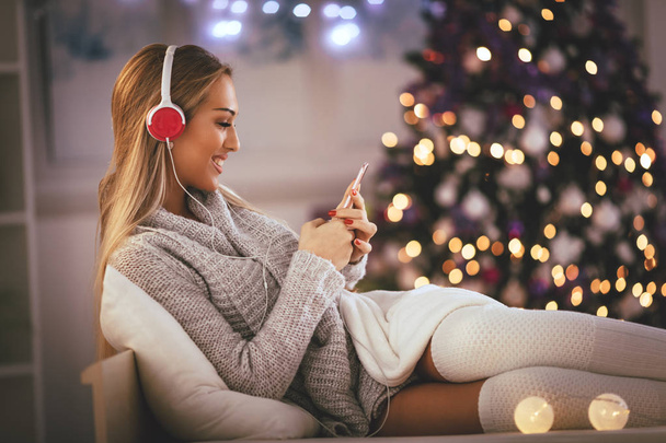 Cute young smiling woman listening music from smartphone during cozy Xmas holidays at home. - 写真・画像
