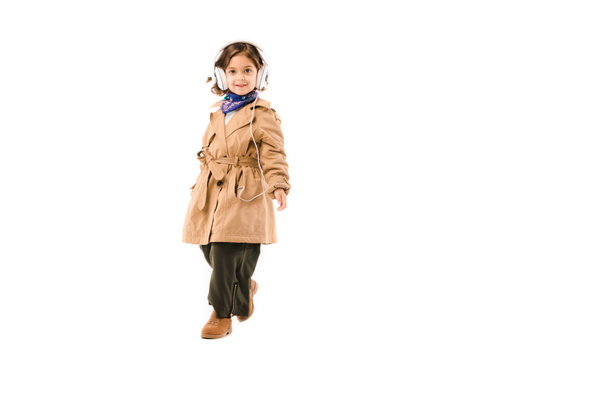 happy little child in stylish trench coat listening music with headphones isolated on white - Фото, изображение
