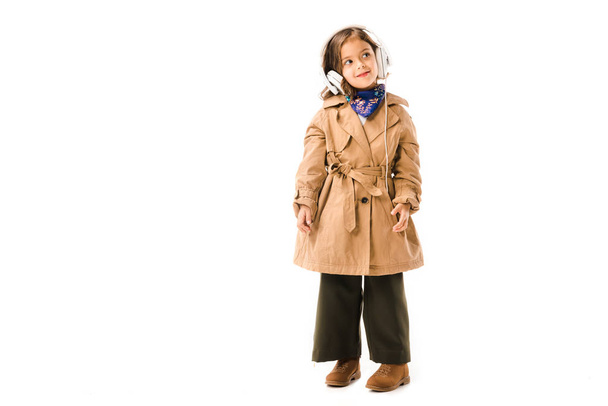 beautiful little child in trench coat listening music with headphones isolated on white - Photo, Image
