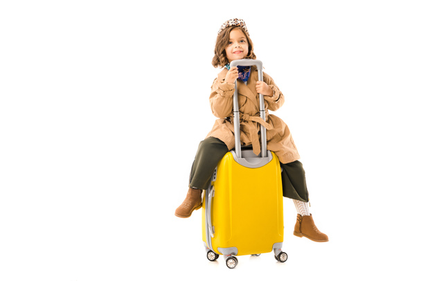 beautiful little child in trench coat sitting on yellow suitcase isolated on white - 写真・画像