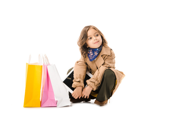 beautiful little child in trench coat sitting on floor with colorful shopping bags isolated on white - Fotó, kép