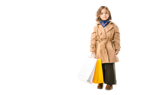 entzückendes stilvolles Kind im Trenchcoat mit bunten Einkaufstaschen, das isoliert auf weiß in die Kamera blickt - Foto, Bild