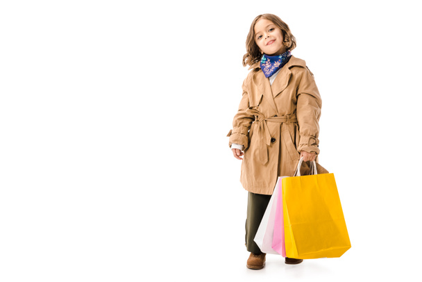adorable petit enfant en trench coat avec des sacs à provisions colorés regardant la caméra isolée sur blanc
 - Photo, image