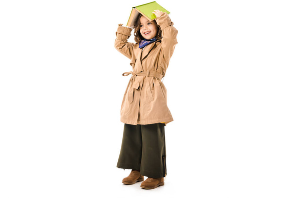 beautiful little child in trench coat covering head with book isolated on white - Photo, Image