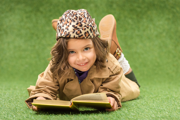 elegante niño sonriente con libro mirando a la cámara mientras está acostado en la hierba verde
 - Foto, imagen