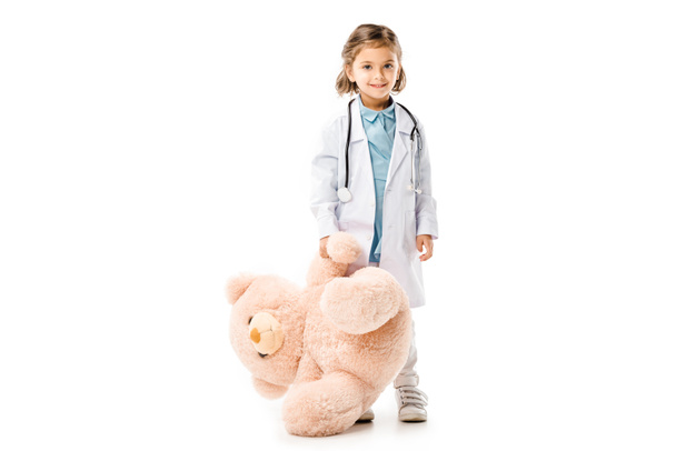dzieciak ubrany w lekarzy biały płaszcz z stetoskop trzyma wielkiego pluszowego misia na białym tle - Zdjęcie, obraz
