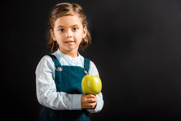 portrait of little schoolgirl with apple in hands looking at camera isolated on black - Zdjęcie, obraz