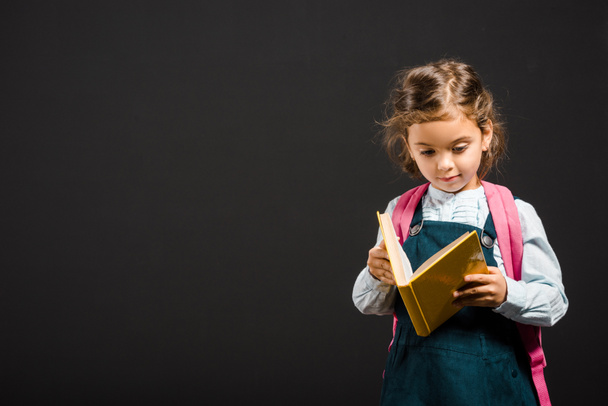 cute schoolgirl with backpack and book in hands isolated on black - 写真・画像