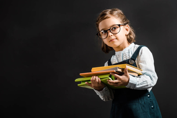 portrait of adorable schoolchild in eyeglasses holding books isolated on black - Photo, image