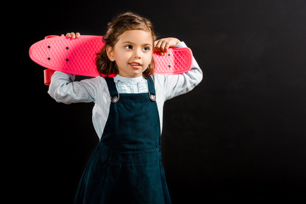 portrait of cute schoolchild holding pink penny board isolated on black - Фото, изображение