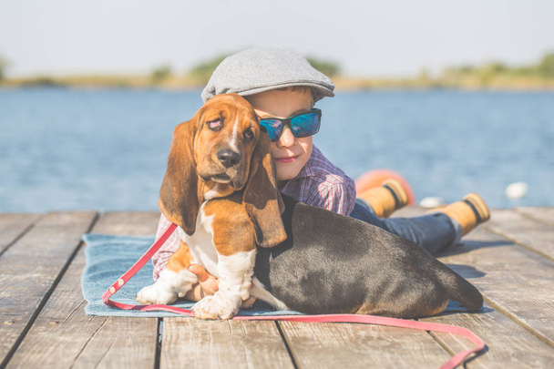 Little cute chłopiec leży nad rzeką z psem. Cieszą się razem w piękny słoneczny dzień. Chłopiec przytulanie jego szczeniak. Dorastając, miłości do zwierząt - psów, czas wolny, Podróże, wakacje. Miejsce - Zdjęcie, obraz