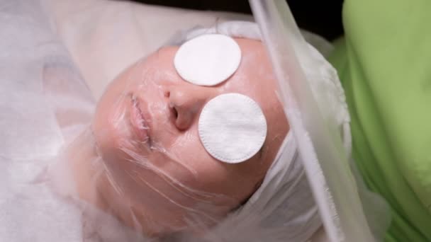 Χέρια των αισθητικός ή έναν γιατρό σε λευκά γάντια καλύπτουν το πρόσωπο του ένα ασιατικό κορίτσι με μια διαφανή μεμβράνη πριν από τη διαδικασία του δέρματος στον ατμό. Μαυρισμένο ανατολίτικο γυναίκα σε ένα γραφείο ιατρικής κοσμετολογίας - Πλάνα, βίντεο