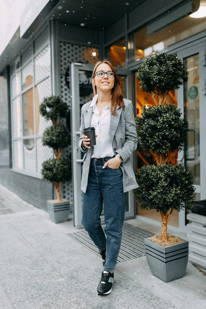 Blogger chica con café de pie en la calle, esperando una reunión. Chica elegante y sonriente. Un comprador feliz, una cafetería moderna
. - Foto, Imagen