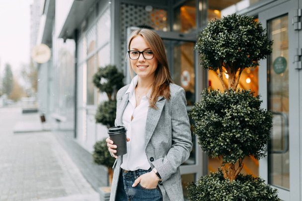 Девушка-блоггер с кофе стоит на улице и ждет встречи. Стильная и улыбающаяся девушка. Счастливый покупатель, современное кафе
. - Фото, изображение