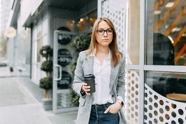 Girl blogger with coffee standing on the street, waiting for a meeting. Stylish and smiling girl. A happy buyer, a modern coffee shop. - Photo, Image