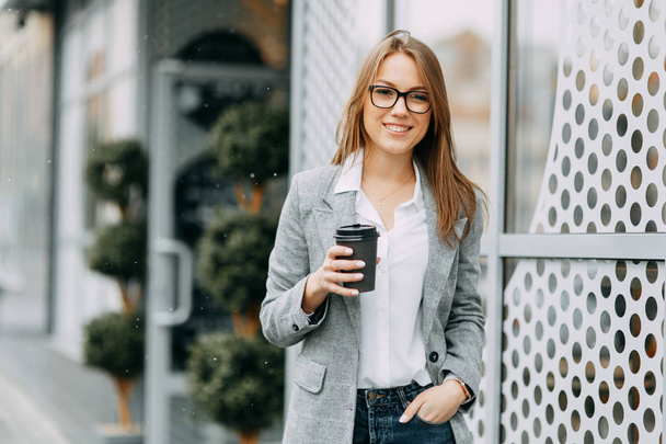 Girl blogger with coffee standing on the street, waiting for a meeting. Stylish and smiling girl. A happy buyer, a modern coffee shop. - Foto, Imagem