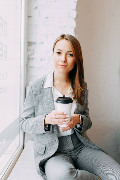 Young blogger girl with coffee sitting and working in a cafe. Bright portraits in the coffee shop, smiles and happy eyes. Business woman, strict style - 写真・画像