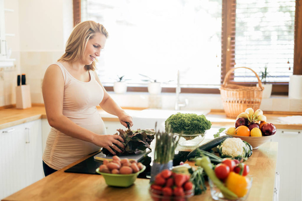 Beautiful pregnant woman preparing meal on kitchen island from fresh ingredients - Foto, imagen