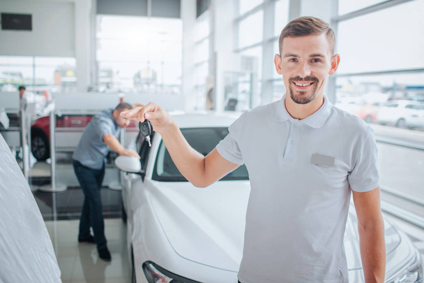 Picture of car deeler stands and looks on camera. He smiles and holds car key. Potential purchaser stands and leans on white car. He looks inside of it. with seriousness. They are in dealing store. - 写真・画像