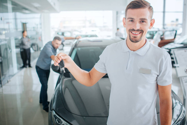 Young and bearded consultant stands and holds key from black car. He looks on caera and smiles. Guy wears white shirt. Potential purchaser is nearby. He looks inside of car. - Фото, изображение