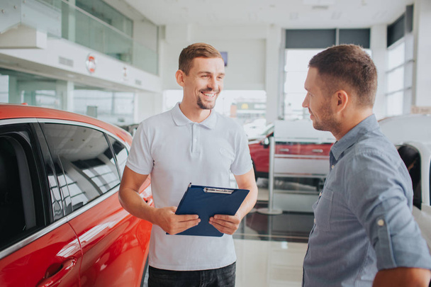 Picture of customer and seller stand together in front of beautiful red car. They look at each other and smile. Seller holds plastic tablet. They are on salon. - Φωτογραφία, εικόνα