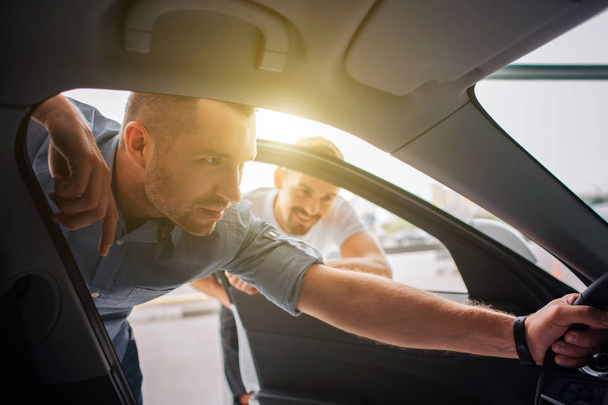 Handsome and confident customer stands and leans to car. He holds steering wheel with one hand. Guy is attentive to details. Manager stands behind him and lean to car. He is smiling and watching. - Photo, image