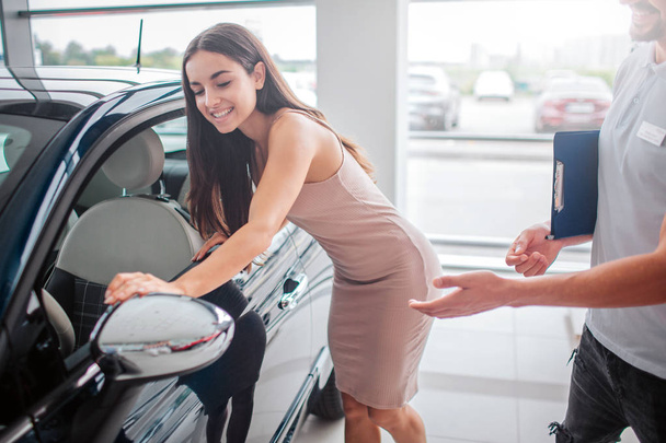 Cheerful and positive young woman looks in car mirror and smiles. She touches it. Attractive young woman likes this car. Consultant stand besides her and points on car. - Foto, immagini