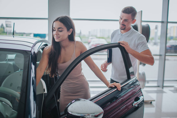 Picture of young woman sit down in car. She touches it and looks with pleasure. Consultant stands behind her. He holds hand on door. Young man looks at woman. - Foto, afbeelding