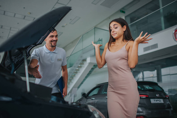 Distracted and lost young woman stands at car with opened front body and keeps hands up aside of her body. She looks down. Consultant stands besides her and smiles. He looks at model. - Foto, immagini