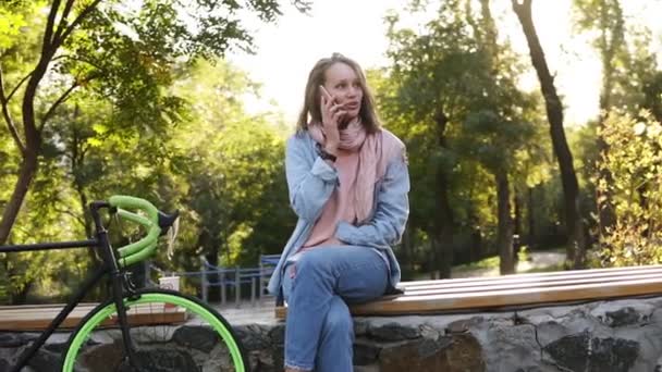Beautiful fair haired girl sitting on the bench in the city park with her trekking bike next to her. Talking by her mobile phone, smiling. Wearing bright pink and blue coloured casual clothes. Trees - Filmati, video
