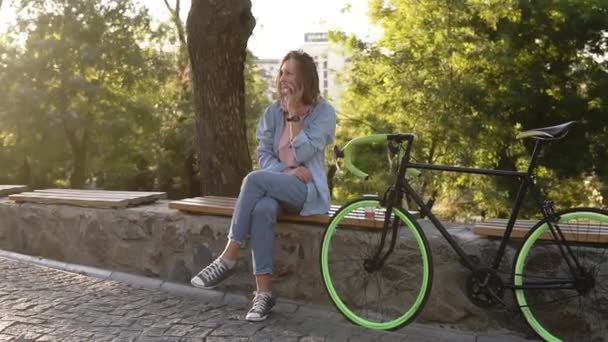 Pretty girl sitting on the bench or parapet in the city park with her trekking bike next to her. Talking by her mobile phone, smiling. Wearing bright pink and blue coloured casual clothes and sneakers - Imágenes, Vídeo