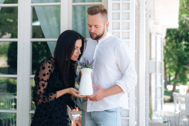 An attractive pair of Europeans rest in their country house. The man brought his beloved flowers from the garden in the vase. The girl gently smells a bouquet of flowers. Lovers spend time together. - Photo, image