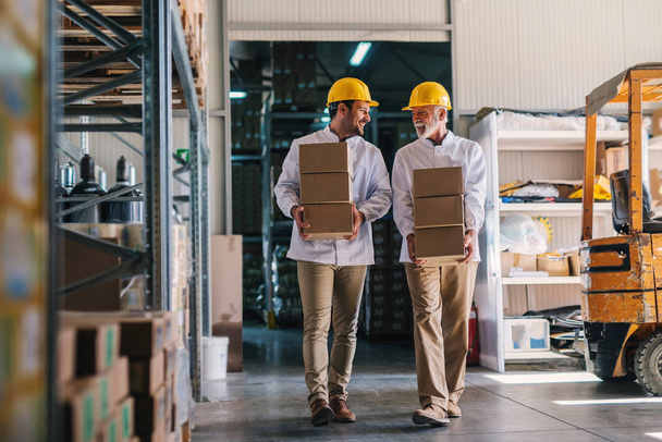 Picture of two male warehouse workers with helmets on their heads carrying boxes in their hands. Talking and walking. - Photo, Image