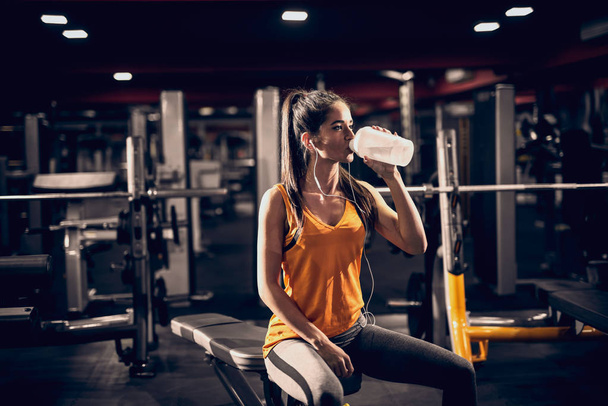 Mujer bebiendo agua con auriculares en los oídos mientras está sentada en el banco. Retroiluminación, gimnasio interior
. - Foto, Imagen