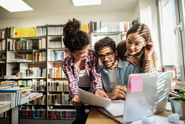  три молодых трудолюбивых студента учатся в библиотеке с помощью ноутбука
 - Фото, изображение