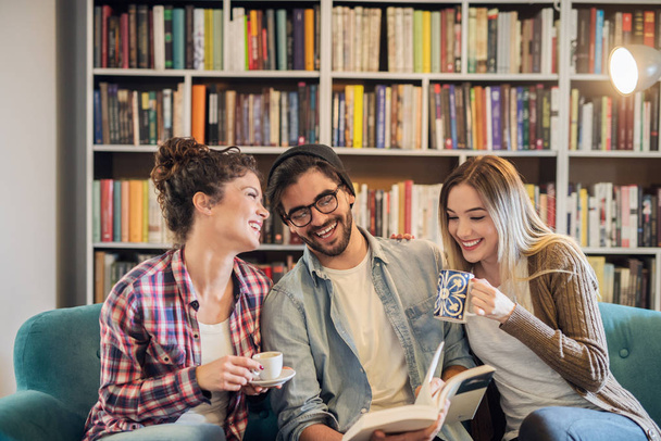 Conceito de educação, biblioteca, estudantes e trabalho em equipe. Três jovens amigos felizes lendo livros sentados na biblioteca e estudando
   - Foto, Imagem