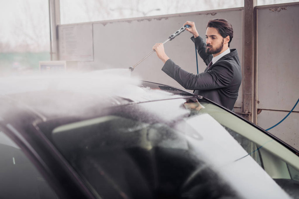 handsome stylish young bearded man in suit washing car at the manual car washing self-service station - Φωτογραφία, εικόνα