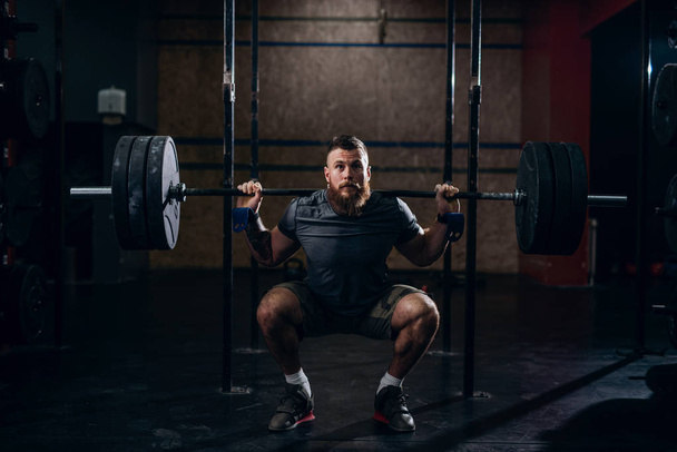 Musculoso caucásico barbudo hombre levantando pesas y haciendo back squat en gimnasio
. - Foto, imagen