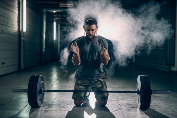 bearded strong bodybuilder preparing to exercise with heavy barbell in gym, using powder - Photo, Image