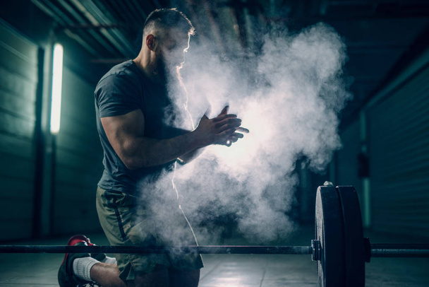 bearded strong bodybuilder preparing to exercise with heavy barbell in gym, using powder - Photo, Image