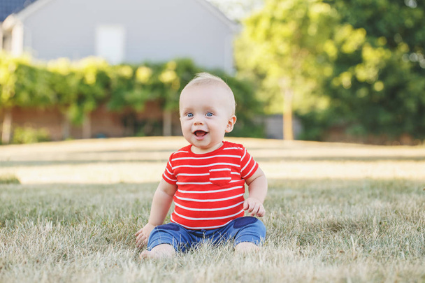 Cute adorable little Caucasian baby boy in red t-shirt and pants sitting in field meadow outside. Little happy smiling child in summer park on sunset. Lovely charming male infant having fun. - Photo, image