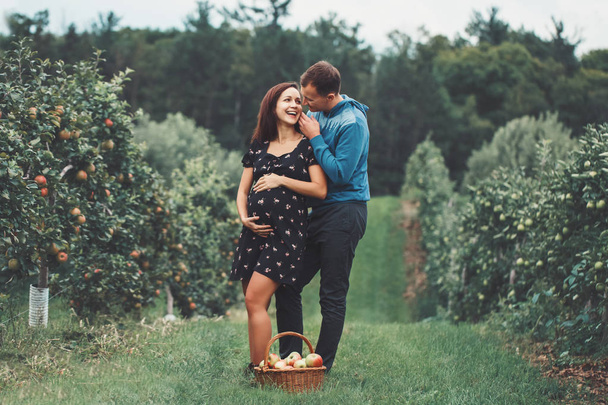 Happy healthy pregnancy and parenting. Portrait of pregnant young brunette Caucasian woman with husband on apple farm. Beautiful expecting mother and future father at countryside, rustic style - Photo, Image