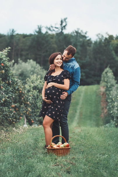Happy healthy pregnancy and parenting. Portrait of pregnant young brunette Caucasian woman with husband on apple farm. Beautiful expecting mother and future father at countryside, rustic style - Photo, Image