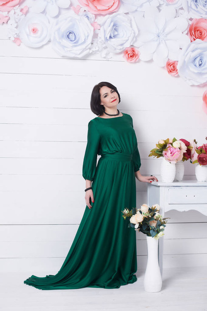 όμορφη μελαχρινή γυναίκα με το πράσινο φόρεμα που ποζάρει με λουλούδια σε ανοιχτόχρωμο φόντο - Φωτογραφία, εικόνα