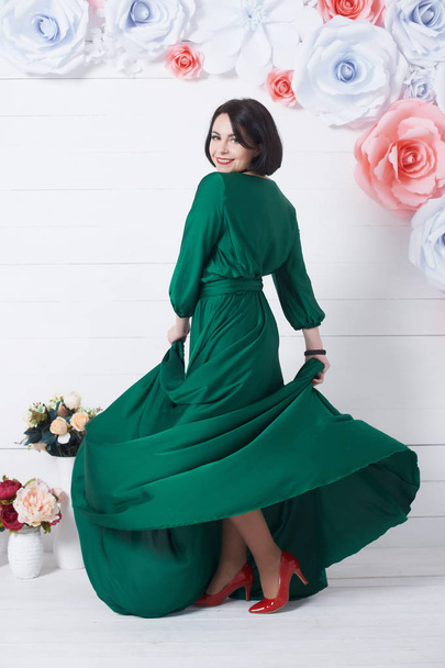 όμορφη μελαχρινή γυναίκα με το πράσινο φόρεμα που ποζάρει με λουλούδια σε ανοιχτόχρωμο φόντο - Φωτογραφία, εικόνα