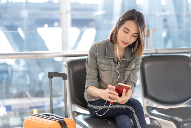 Happy young Asian woman listening music with headphones and mobile phone in terminal hall while waiting for her flight - Valokuva, kuva